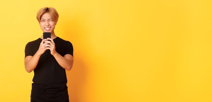 Stylish handsome asian guy taking picture on smartphone and smiling, photographing with mobile phone, standing yellow background.