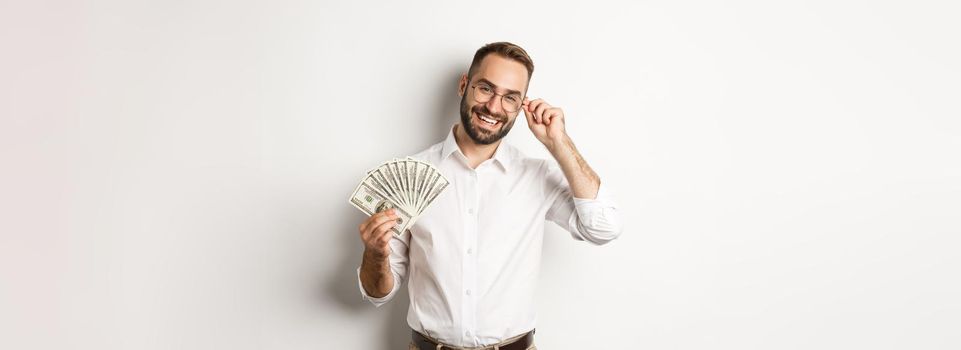 Handsome successful businessman holding money, fixing glasses on nose, standing over white background.