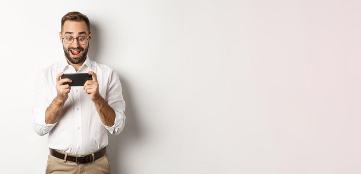 Man looking excited and surprised at mobile phone, holding smartphone horizontally, standing over white background.