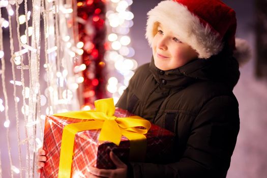 Happy child in a red Santa Claus hat with wrapped Christmas gift near shining light bulbs in winter evening. The concept of Christmas and New Year.