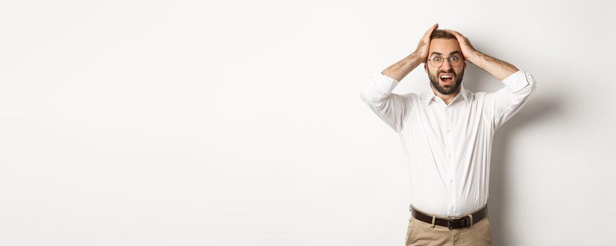 Frustrated businessman holding hands on head, looking shocked and anxious, standing over white background.
