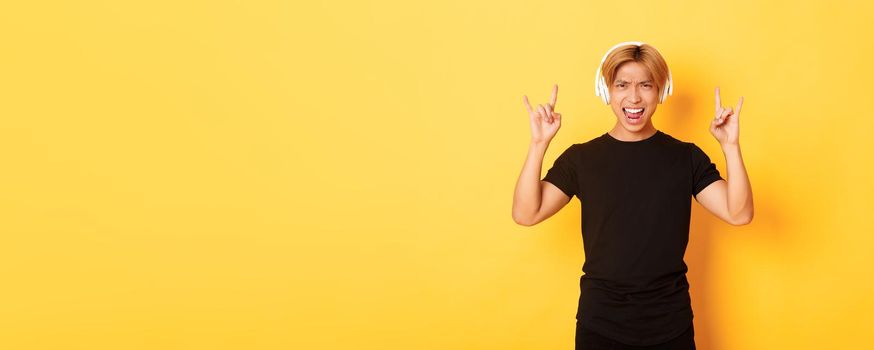 Joyful handsome asian guy enjoying listening music in headphones, showing rock-n-roll gesture, standing over yellow background.