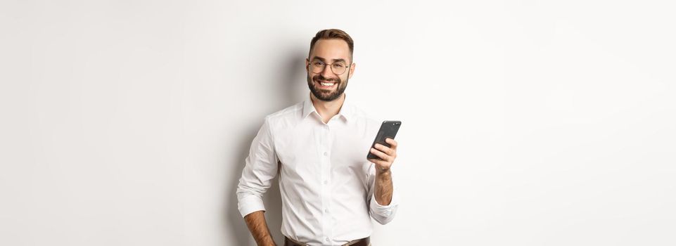 Handsome manager using smartphone and smiling pleased, sending text message, standing over white background.