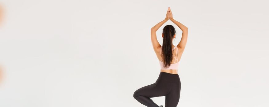 Sport, gym and healthy body concept. Full length rear view of slim brunette asian girl in active wear practice yoga, workout alone, standing with hands clasped over head in asana, meditating.
