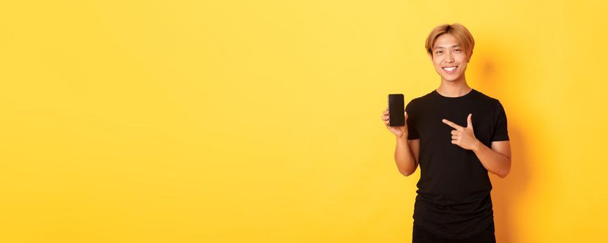 Portrait of handsome asian man showing something on smartphone screen, pointing at mobile phone display, standing over yellow background.