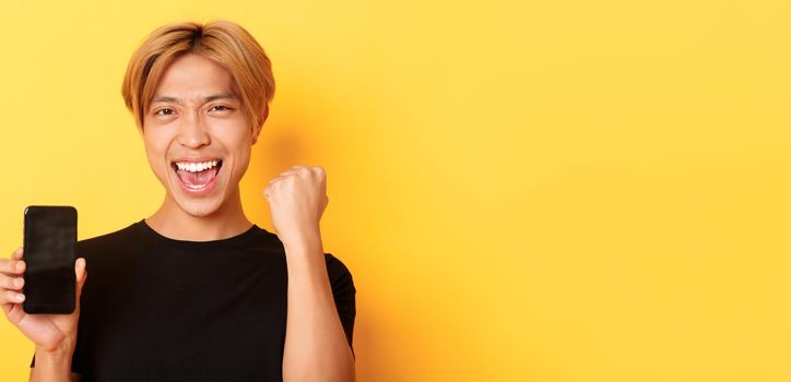 Close-up of happy rejoicing asian guy showing smartphone screen and saying yes, fist pump as triumphing, winning or achieve goal, yellow background.