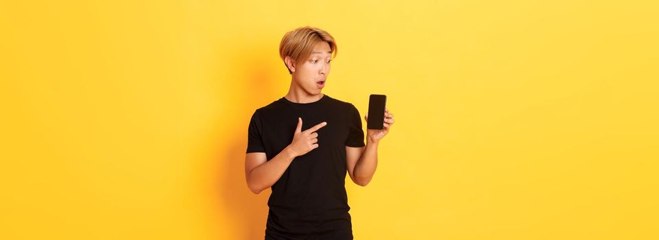 Portrait of handsome asian man showing something on smartphone screen, pointing at mobile phone display, standing over yellow background.