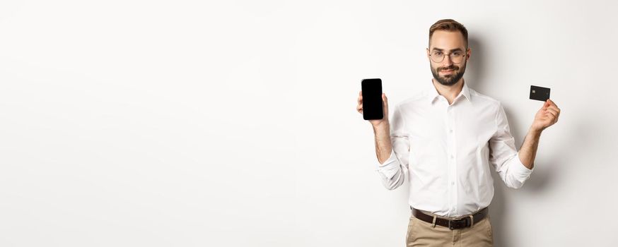 Handsome bearded man showing mobile phone and credit card, shopping online, standing over white background.
