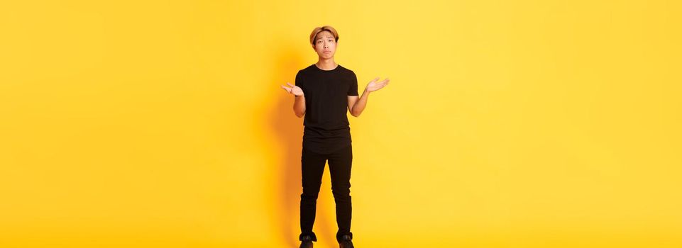 Full-length of disappointed and frustrated asian guy shrugging, looking sad, yellow background.