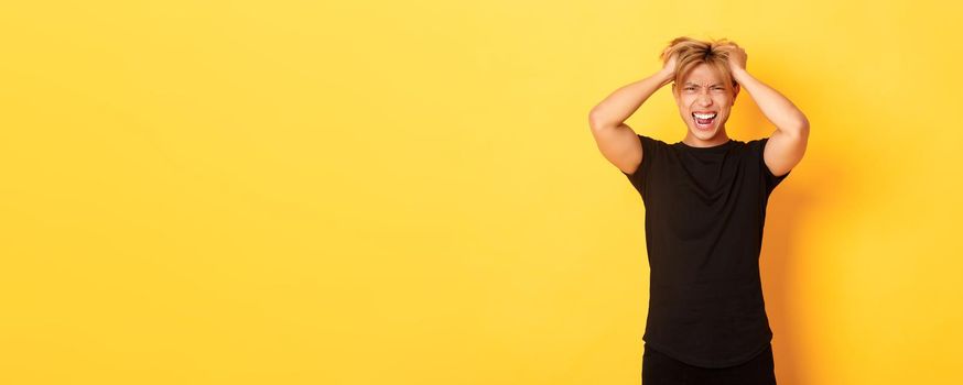 Portrait of pissed-off angry asian man tossing haircut and yelling furious, standing over yellow background.