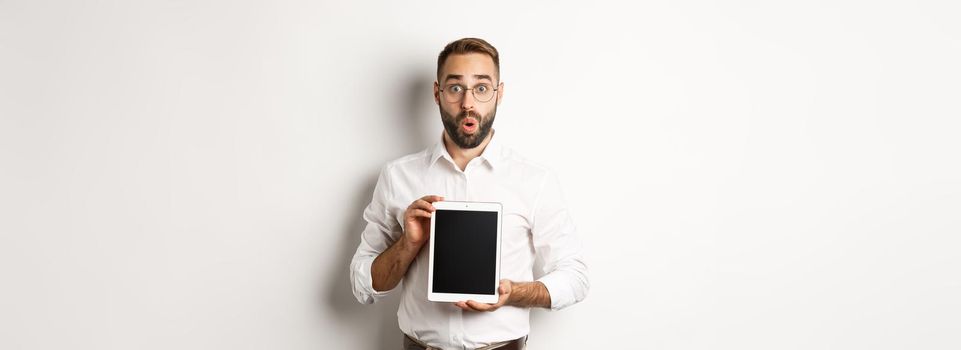 Surprised man in glasses, showing digital tablet screen, looking amazed, standing over white background.