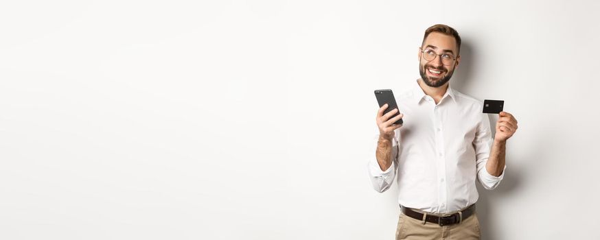 Business and online payment. Image of handsome man thinking while holding credit card and smartphone, standing against white background.