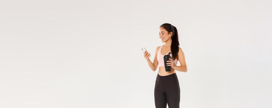 Full length of smiling fit and healthy female fitness coach, asian sportswoman checking water tracker reminder in smartphone, using application during workout in gym, white background.