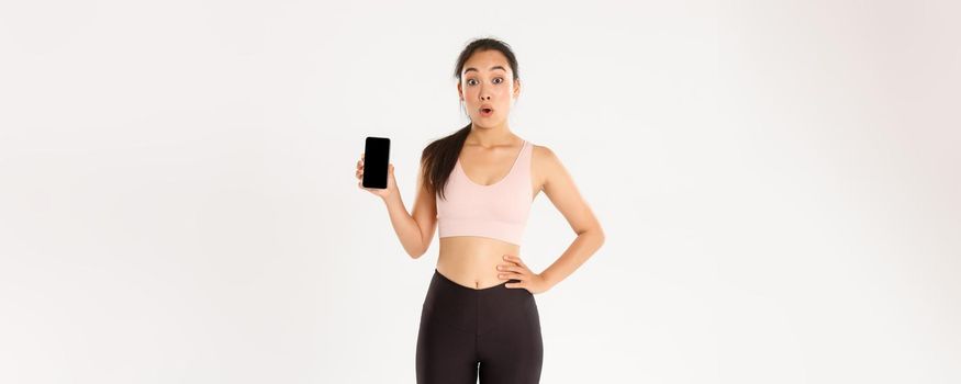 Sport, technology and active lifestyle concept. Amazed and impressed asian sportswoman, athlete say wow look thrilled and showing mobile phone fitness application, workout tracker app.