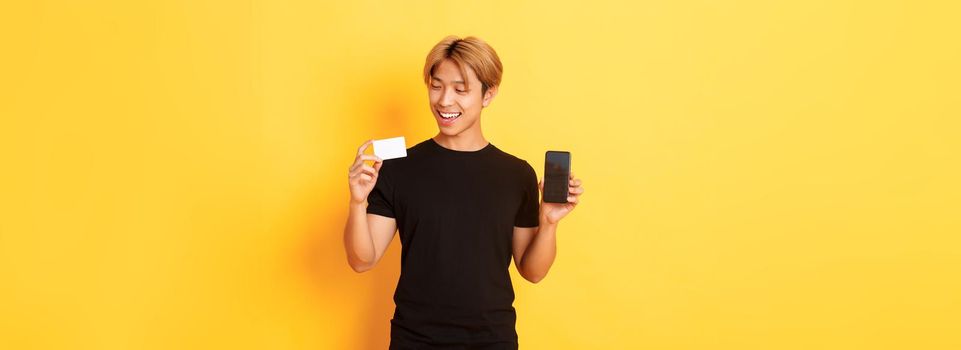 Handsome satisfied asian guy holding credit card and showing smartphone screen, smiling pleased, standing yellow background.