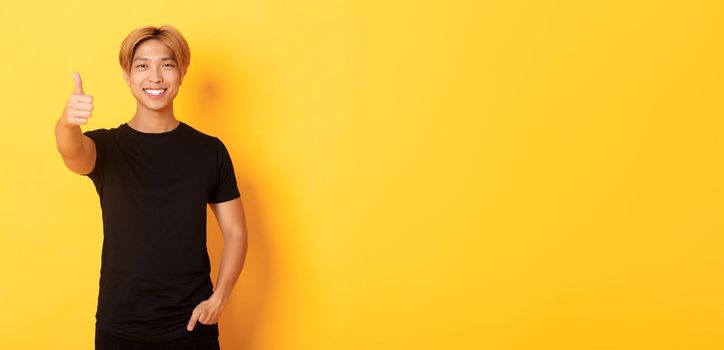 Portrait of satisfied asian man with blond hair, standing over yellow background and showing thumbs-up.