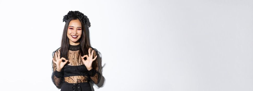 Cheerful beautiful asian woman in witch dress showing okay gestures and smiling satisfied, approve halloween costume or advertisement, standing over white background.