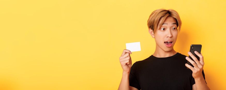 Close-up of amazed asian guy looking at mobile phone while shopping online and showing credit card, yellow background.