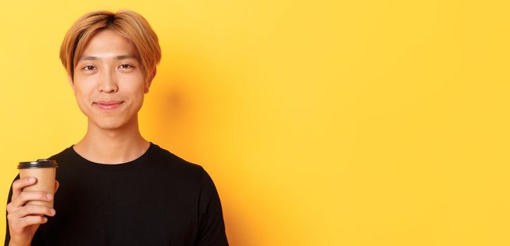 Close-up of handsome blond asian man drinking coffee and smiling pleased, standing over yellow background.