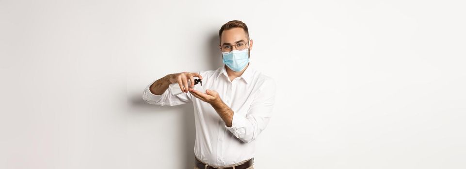Covid-19, social distancing and quarantine concept. Office worker in medical mask clean hands with antiseptic, using sanitizer, standing over white background.