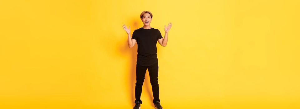 Full-length of surpirsed and excited, smiling blond asian guy raising hands up amazed, yellow background.