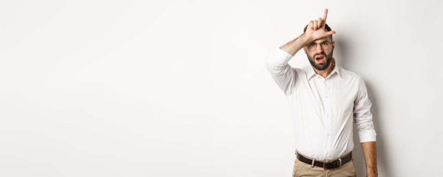 Shocked guy showing loser sign on forehead, complaining, standing over white background.