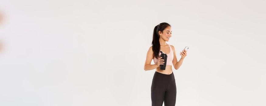 Full length of smiling fit and healthy female fitness coach, asian sportswoman checking water tracker reminder in smartphone, using application during workout in gym, white background.