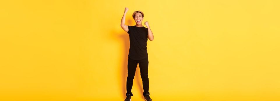 Full-length of triumphing handsome asian man with blond hair, raising hands up in celebration, saying yes, standing yellow background.
