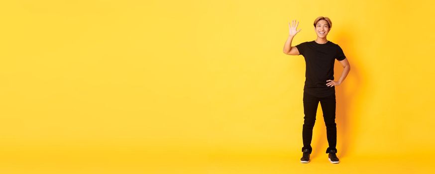 Full-length of friendly handsome asian man waving hand in hello, smiling and saying hi over yellow background.