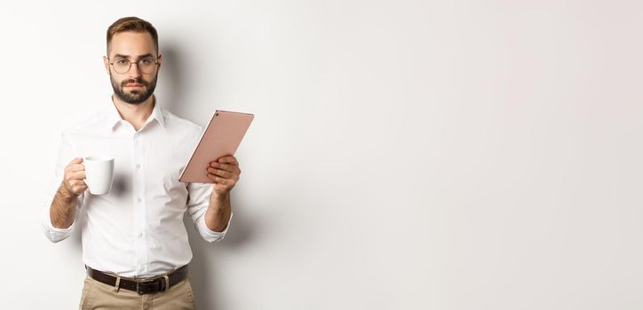 Confident male manager reading work on digital tablet and drinking coffee, standing over white background.