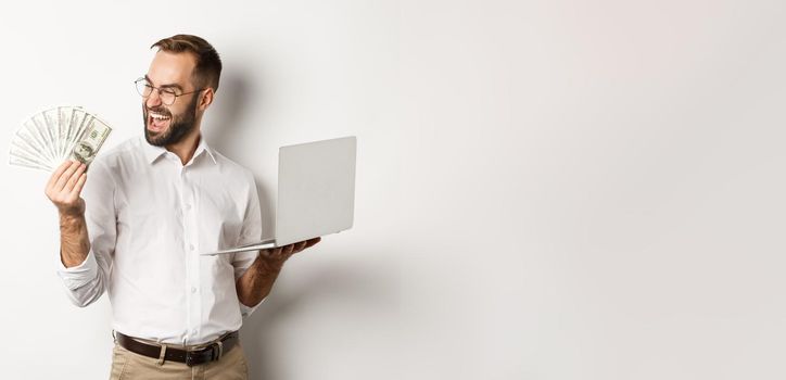 Business and e-commerce. Successful businessman using laptop for work and holding money, standing over white background.