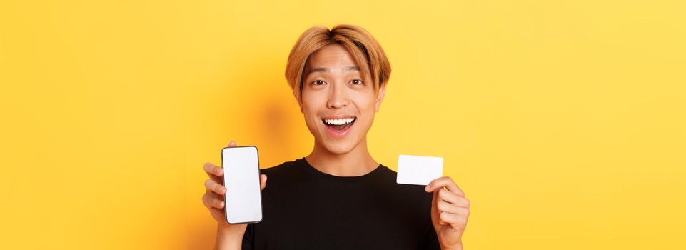 Amazed happy asian guy showing credit card and smartphone screen, smiling fascinated, standing yellow background.