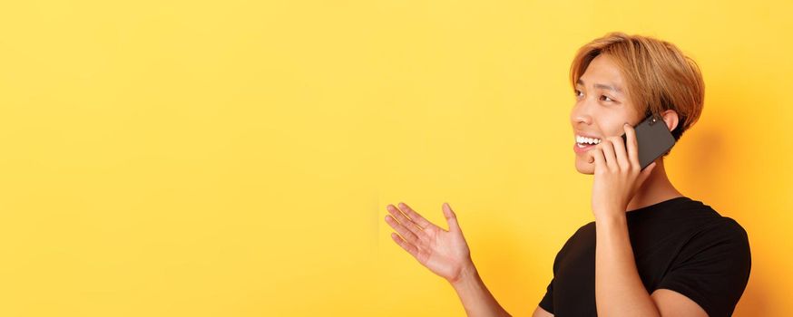 Close-up of attractive korean guy smiling and talking on mobile phone happy, turning left, standing yellow background.