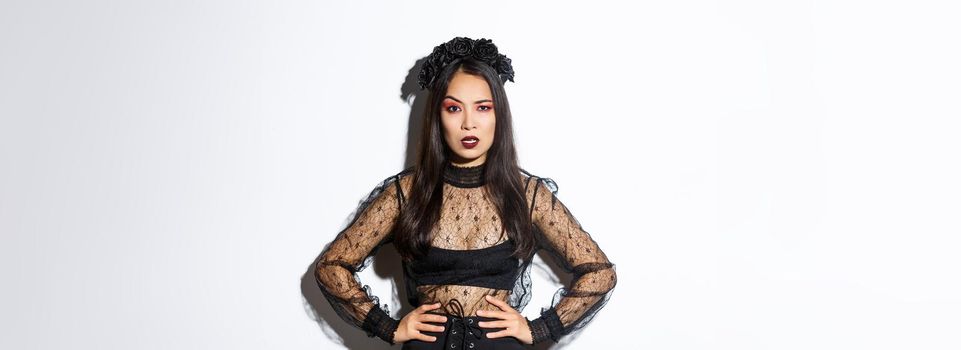 Angry and pissed-off asian female magician, evil witch in black dress and wreath looking mad at someone, frowning and looking judgemental, standing over white background.
