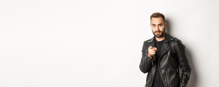Handsome and cool bearded guy pointing finger at camera, wearing leather jacket, standing sassy against white background.