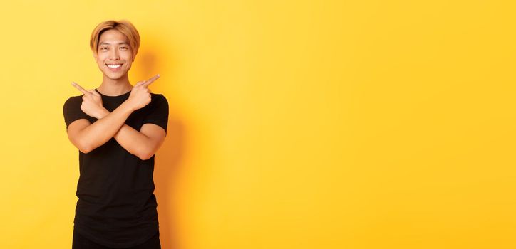 Portrait of handsome determined blond asian guy, student pointing fingers sideways, showing two variants, standing yellow background.
