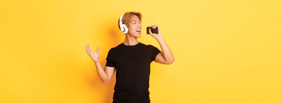 Joyful attractive korean guy in headphones, playing karaoke app, singing into mobile phone microphone, standing yellow background.