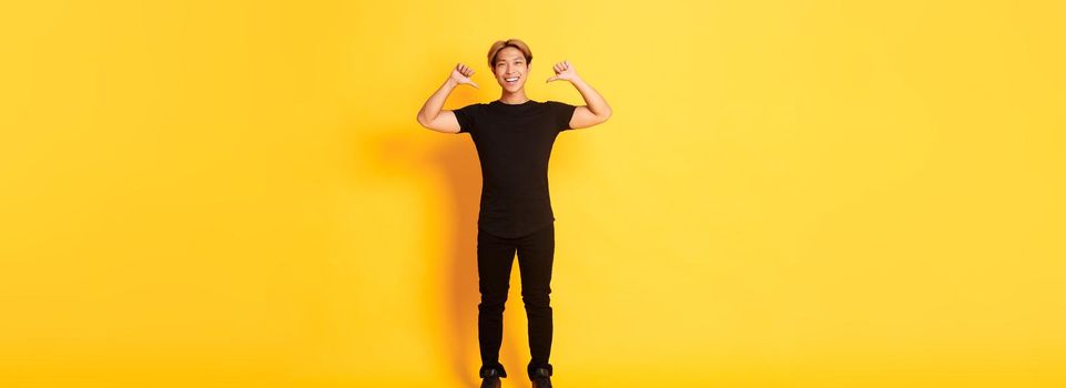 Confident attractive blond asian guy, smiling sassy and pointing at himself, standing over yellow background.