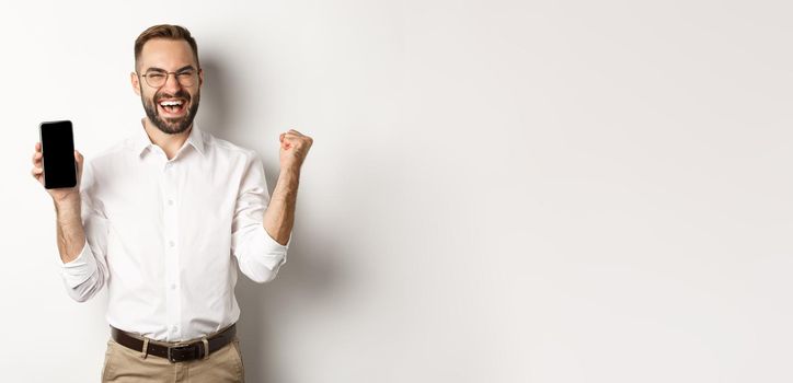 Successful business man showing mobile screen, rejoicing on winning online prize, achieve app goal, standing against white background.