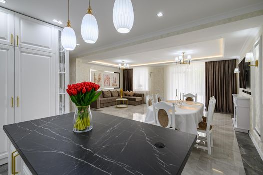 Luxurious white large modern studio interior with kitchen, black marble countertop over kitchen island, dining area with served table