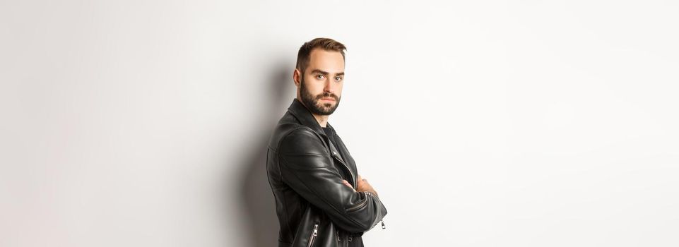 Profile of confident and handsome bearded man in black leather jacket, turn face at camera and looking serious, white background.