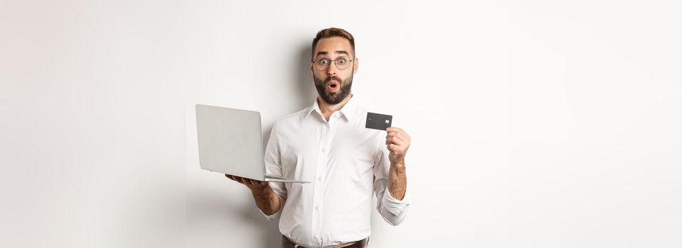 Online shopping. Surprised man holding laptop and credit card, shop internet store, standing over white background.