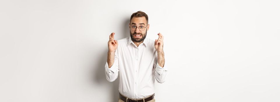 Worried man making a wish, cross fingers and hope for relish, standing over white background.