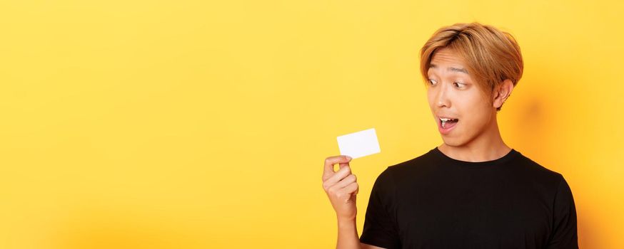 Close-up of amazed asian guy talking about credit card, standing over yellow background.