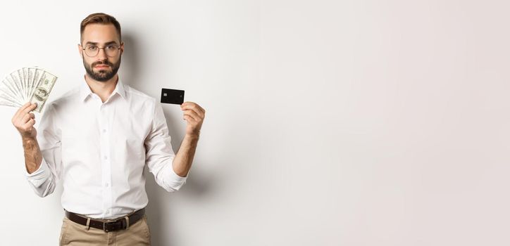 Serious businessman looking at camera, holding credit card and money, standing over white background. Concept of shopping and finance.