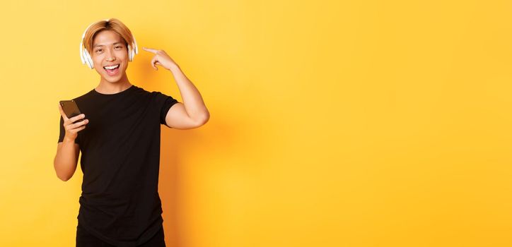 Happy satisfied asian guy likes music or podcast, pointing finger at headphones, holding smartphone, standing yellow background.