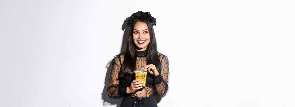 Smiling cute asian woman celebrating halloween, holding sweets and grinning happy, trick or treating in witch costume.