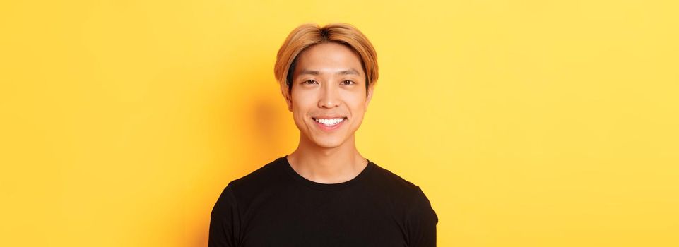 Close-up of handsome blond asian guy in black t-shirt, smiling happy at camera, standing over yellow background.