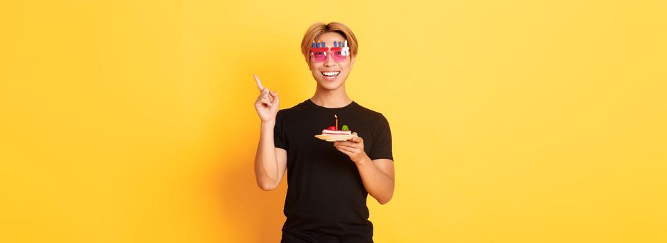 Cheerful, handsome asian birthday guy in funny sunglasses, holding b-day cake with candle and pointing finger upper right corner at your logo, yellow background.