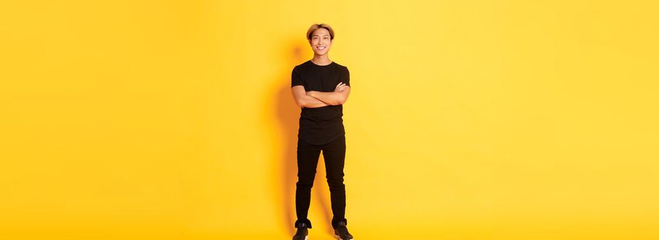 Full length of smiling confident asian blong guy, cross arms chest over yellow background.